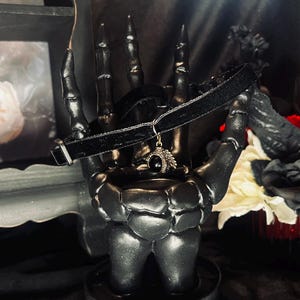 May include: A black velvet choker necklace with a silver pendant featuring a black stone and ornate detailing, displayed on a black hand-shaped sculpture. The background includes a floral arrangement with red, black, and white flowers.