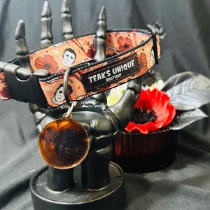 May include: A Halloween-themed dog collar with a peach background featuring images of a masked character and flowers. The collar has a black buckle and a matching tag. The text "TEAKS UNIQUE BOUTIQUE" is visible on the collar.