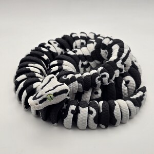 Puede incluir: Escultura de serpiente enroscada en blanco y negro. El cuerpo de la serpiente est&aacute; hecho de segmentos redondeados, con un patr&oacute;n de cebra. La cabeza es blanca con rayas negras y un ojo verde. Sobre un fondo blanco.