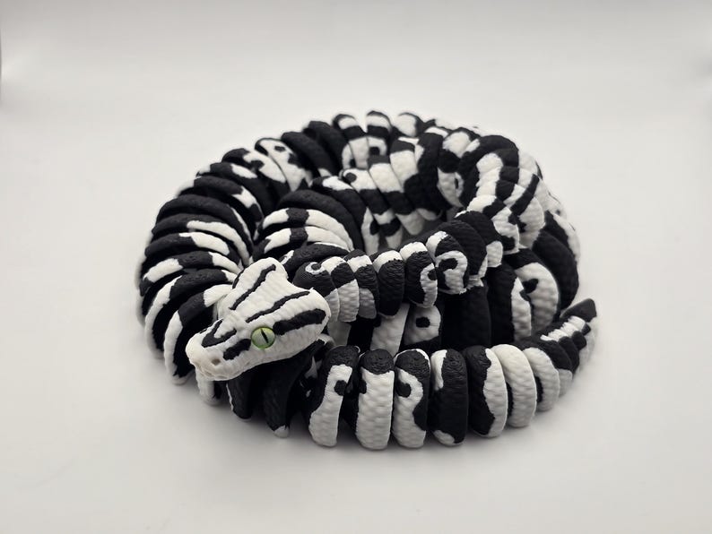 Puede incluir: Escultura de serpiente enroscada en blanco y negro. El cuerpo de la serpiente est&aacute; hecho de segmentos redondeados, con un patr&oacute;n de cebra. La cabeza es blanca con rayas negras y un ojo verde. Sobre un fondo blanco.