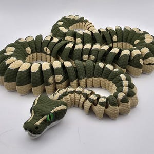 Puede incluir: Escultura detallada de una serpiente en tonos verdes y beige. El cuerpo de la serpiente est&aacute; segmentado, con un patr&oacute;n texturizado. La cabeza presenta ojos verdes y un dise&ntilde;o detallado, creando una apariencia realista. La escultura se exhibe sobre una superficie blanca.