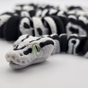 Puede incluir: Primer plano detallado de una figura de serpiente. La serpiente es principalmente blanca con marcas negras y un ojo verde. El cuerpo segmentado est&aacute; enfocado, con la cabeza prominentemente mostrada. El dise&ntilde;o es realista y detallado.