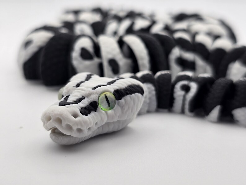 Puede incluir: Primer plano detallado de una figura de serpiente. La serpiente es principalmente blanca con marcas negras y un ojo verde. El cuerpo segmentado est&aacute; enfocado, con la cabeza prominentemente mostrada. El dise&ntilde;o es realista y detallado.