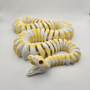 Puede incluir: Un juguete de serpiente articulada blanca y amarilla con la boca abierta y ojos rojos. La serpiente est&aacute; hecha de piezas segmentadas, lo que le permite moverse y enrollarse. El juguete est&aacute; dise&ntilde;ado para parecerse a una pit&oacute;n.
