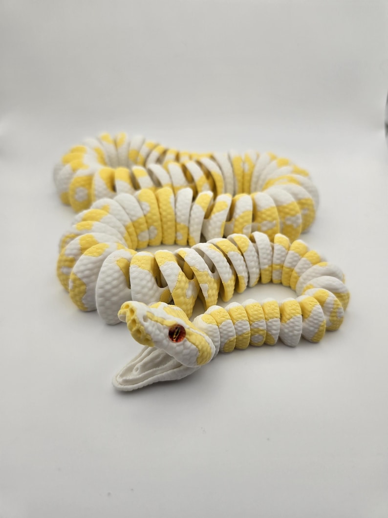 Puede incluir: Un juguete de serpiente articulada blanca y amarilla con la boca abierta y ojos rojos. La serpiente est&aacute; hecha de piezas segmentadas, lo que le permite moverse y enrollarse. El juguete est&aacute; dise&ntilde;ado para parecerse a una pit&oacute;n.