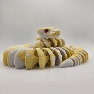 Puede incluir: Un juguete de serpiente articulada amarilla y blanca con ojos rojos. La serpiente est&aacute; hecha de piezas segmentadas, lo que le permite posar en varias posiciones. El juguete est&aacute; dise&ntilde;ado para parecerse a una serpiente de cascabel.