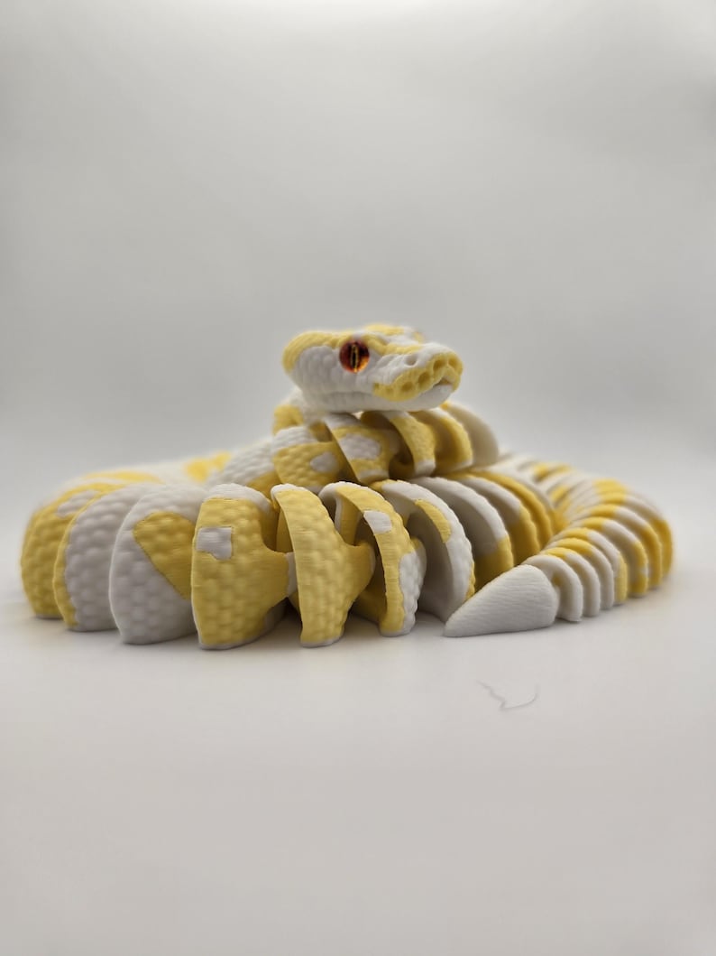 Puede incluir: Un juguete de serpiente articulada amarilla y blanca con ojos rojos. La serpiente est&aacute; hecha de piezas segmentadas, lo que le permite posar en varias posiciones. El juguete est&aacute; dise&ntilde;ado para parecerse a una serpiente de cascabel.