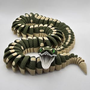 以下が含まれることがあります： 緑とベージュの色合いの、関節式ヘビのおもちゃ。ヘビの頭は口を開けており、緑色の目があります。体はセグメント化され、テクスチャパターンがあります。おもちゃは柔軟でポーズが取れるように設計されています。