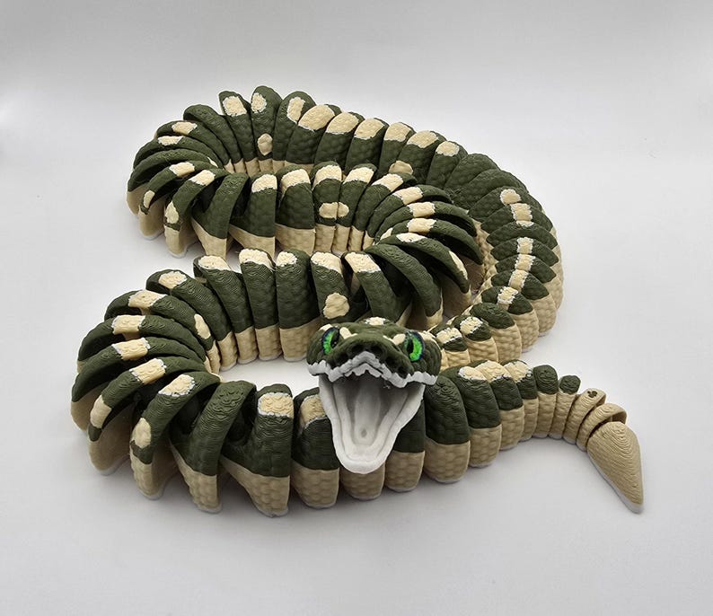 Puede incluir: Un juguete de serpiente articulada en tonos verdes y beige. La cabeza de la serpiente est&aacute; abierta con ojos verdes. El cuerpo est&aacute; segmentado, con un patr&oacute;n texturizado. El juguete est&aacute; dise&ntilde;ado para ser flexible y posable.