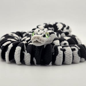 Puede incluir: Figura de serpiente enroscada con un patr&oacute;n en blanco y negro. La cabeza de la serpiente es blanca con ojos verdes. El cuerpo es una mezcla de segmentos negros y blancos, creando un contraste llamativo. La figura est&aacute; sobre un fondo blanco liso.