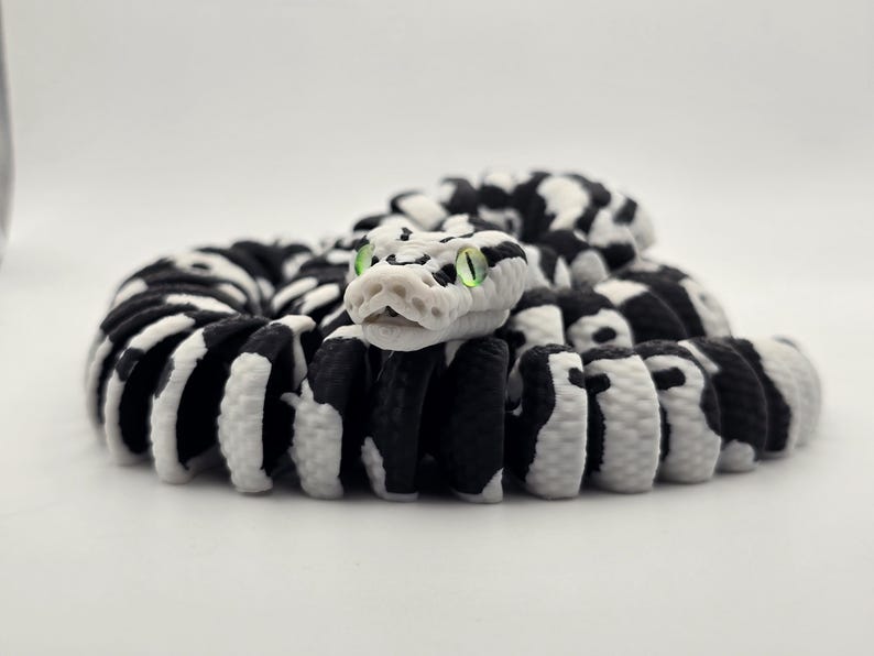 Puede incluir: Figura de serpiente enroscada con un patr&oacute;n en blanco y negro. La cabeza de la serpiente es blanca con ojos verdes. El cuerpo es una mezcla de segmentos negros y blancos, creando un contraste llamativo. La figura est&aacute; sobre un fondo blanco liso.