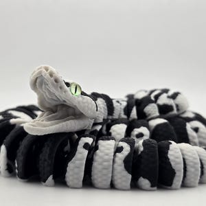 Puede incluir: Escultura detallada de una serpiente con la boca abierta. El cuerpo de la serpiente es negro y blanco, con un patr&oacute;n de rayas y manchas alternas. La cabeza de la serpiente es blanca, con una textura detallada y un ojo verde.