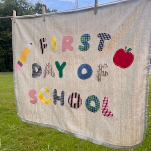 Könnte beinhalten: Ein beigefarbenes Stoffbanner mit dem applizierten Text FIRST DAY OF SCHOOL aus verschiedenen bunten Karostoffen und Mustern. Das Banner enthält eine gelbe Bleistift-Applikation auf der linken und eine rote Apfel-Applikation auf der rechten Seite.