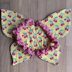 May include: Butterfly wings made of yellow fabric with a cherry pattern. The wings have a red and white gingham ruffle trim. The wings are designed to be worn by a child.
