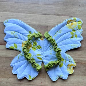 May include: Quilted butterfly wings in light blue fabric with a yellow floral design and green trim. The wings are designed to be worn, featuring a green, ruffled neckband. The wings are made of fabric.