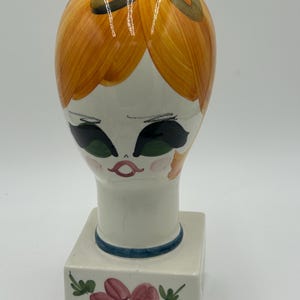 Puede incluir: Un expositor de cerámica en forma de cabeza con base blanca, cabello naranja pintado, un lazo marrón, ojos verdes grandes y mejillas rosadas. La base cuadrada está decorada con una flor rosa pintada a mano y hojas verdes en el frente.