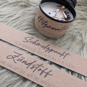 Könnte beinhalten: Eine Kerze in einer schwarzen Dose mit einem braunen Etikett mit deutschem Text. Die Kerze hat ein weißes Wachs mit goldenen, schwarzen und transparenten dekorativen Elementen. Zwei braune Papierstreifen mit deutschem Text befinden sich im Vordergrund.