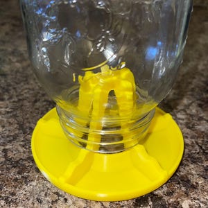 May include: A clear glass jar with a yellow plastic base and a yellow insert. The jar has a wide mouth and a textured surface. The yellow base is round with a raised edge.