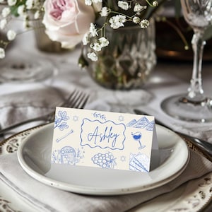 May include: A white place card with blue illustrations of a Star of David, a wine glass, a grapevine, and a matzah. The name "Ashley" is written in blue cursive.