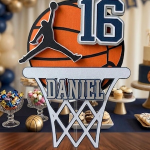 May include: A basketball-themed cake topper with a basketball hoop design. The topper features the name "DANIEL" and the number "16" in blue and silver. A silhouette of a basketball player is also included. The background includes party decorations.