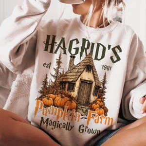 Könnte beinhalten: Cremefarbenes Sweatshirt mit einer Grafik eines Häuschens, Kürbissen und dem Text "Hagrid's Pumpkin Farm Magically Grown". Das Design beinhaltet das Jahr "1981" und ein rustikales Herbstthema.