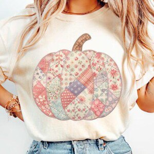 May include: A cream-coloured t-shirt with a patchwork pumpkin design. The pumpkin is made up of various floral and patterned fabrics in pinks, purples, and greens. The shirt has short sleeves and is worn with blue jeans.