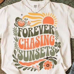 Könnte beinhalten: Cremefarbenes T-Shirt mit dem Text "FOREVER CHASING SUNSETS" in Orange und Grün, mit einer Sonnengrafik. Das Shirt hat einen Rundhalsausschnitt und kurze Ärmel. Eine Halskette ist am Ausschnitt sichtbar.