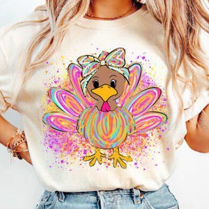 May include: A cream-colored t-shirt featuring a colorful turkey graphic. The turkey has a brown head, yellow beak, and rainbow-colored feathers and bow. The design is surrounded by a splash of pink, purple, and yellow paint.