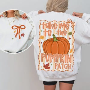 Peut inclure: Sweat-shirt blanc avec un motif sur le thème de l'automne. Le dos présente une citrouille orange avec le texte "Take Me To The Pumpkin Patch". L'avant a un motif de nœud et de feuilles. Vêtement d'automne confortable.