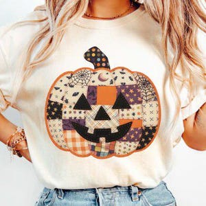 May include: Cream-colored t-shirt featuring a patchwork pumpkin design. The pumpkin has a black jack-o'-lantern face and is made of various patterned fabrics, including orange, purple, and black. The design is centered on the shirt, perfect for Halloween.