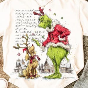Könnte beinhalten: Weißes T-Shirt mit einer Illustration von Grinch und Max. Der Grinch trägt einen roten Mantel mit weißem Besatz, und Max trägt einen roten Schal und Geweih. Der Text auf dem Shirt lautet: "Max never needed words..."