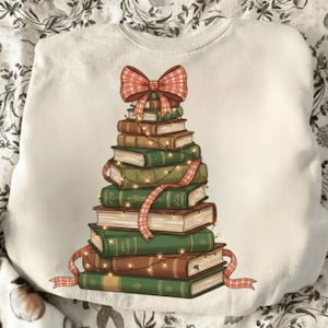 Puede incluir: Sudadera blanca con un diseño de árbol de Navidad hecho con libros apilados. Los libros son marrones y verdes, decorados con un lazo y una cinta a cuadros rojos, y luces de hadas. Una prenda festiva.