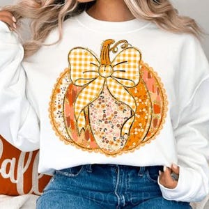 May include: White sweatshirt featuring a pumpkin design with a gingham bow and floral accents. The pumpkin is in shades of orange and peach, with a decorative lace trim. A fall-themed apparel item.