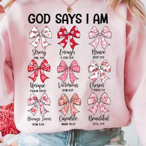 Pode incluir: Camisola rosa claro com o texto "GOD SAYS I AM" acima de nove desenhos de laços. Cada laço tem um design diferente e está emparelhado com uma palavra como "Forte" e uma referência bíblica.