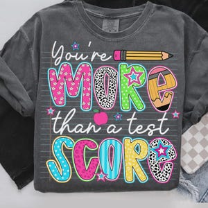 Op de afbeelding: Donkergrijze sweatshirt met de kleurrijke en speelse tekst "You're MORE than a test SCORE". Het ontwerp bevat een potlood, sterren en een appel, perfect voor leraren of leerlingen.