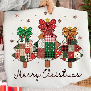 Peut inclure: Tissu blanc avec trois sapins de Noël en patchwork, chacun surmonté d'un nœud et d'une étoile. Les arbres sont dans les tons de vert, rouge et marron. L'inscription "Merry Christmas" est écrite en dessous.