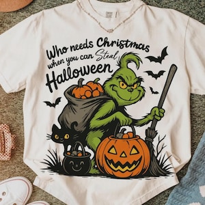 以下が含まれることがあります： ハロウィーンをテーマにしたグラフィックが施されたオフホワイトのTシャツ。デザインは、ほうきとジャック・オー・ランタンを持った緑色のキャラクターと、「Who needs Christmas when you can steal Halloween」というテキストが特徴です。コウモリと黒猫も含まれています。