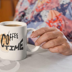 Puede incluir: Taza de café de cerámica blanca con las palabras "Coffee Time" en letras negras y una ilustración de café helado. La taza está llena de café y la sostiene una persona.