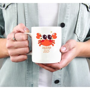 Op de afbeelding: Witte keramische mok met een cartoon krab ontwerp in oranje, rood en zwart. De mok heeft de woorden "SUMMER 2025" in oranje gedrukt. De mok wordt door een persoon vastgehouden.