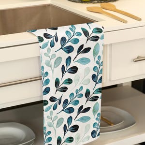 Puede incluir: Paño de cocina blanco con un patrón repetido de hojas verde azulado y azul oscuro. Las hojas tienen un efecto acuarela y están dispuestas sobre un fondo blanco. La toalla está colgada sobre un armario blanco.