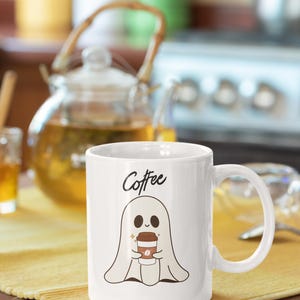 Puede incluir: Taza de café de cerámica blanca con una ilustración de un fantasma sosteniendo una taza de café. La palabra "Coffee" está escrita encima del fantasma. La taza tiene un asa curva y está sobre una superficie amarilla.