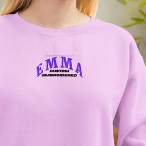 Puede incluir: Una sudadera lila con el texto bordado "EMMA CUSTOM EMBROIDERED" en morado y negro. La prenda es de color liso y parece estar hecha de una tela suave.