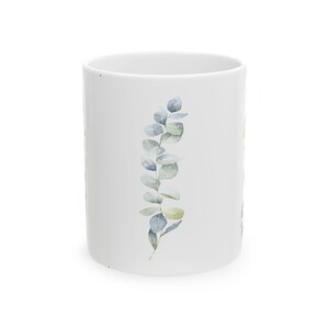Puede incluir: Taza de cerámica blanca con un diseño de rama de eucalipto en acuarela. Las hojas son una mezcla de azul y verde, creando una estética natural. La taza es cilíndrica y adecuada para bebidas.