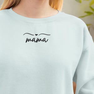 Puede incluir: Sudadera azul claro con la palabra "mama" en escritura cursiva negra, con un pequeño corazón encima. La sudadera tiene un diseño sencillo y probablemente está hecha de una tela suave. El estilo general es informal.