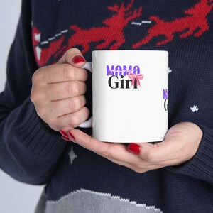 Puede incluir: Taza de cerámica blanca con las palabras "Mama Girl" en morado y negro, con un lazo rosa. La taza es sostenida por una persona que lleva un suéter azul marino con un estampado de renos rojos.
