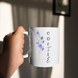 Puede incluir: Taza de café de cerámica blanca con la palabra "COFFEE" en escritura negra. La taza presenta una ilustración de acuarela de flores de campanilla. La taza está sostenida por una persona.