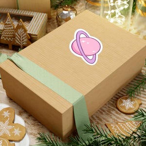 Puede incluir: Una pegatina con un planeta rosa con un anillo morado y estrellas blancas. La pegatina está colocada sobre una caja marrón con una cinta verde claro. La caja está rodeada de adornos navideños, incluyendo galletas de jengibre y agujas de pino.