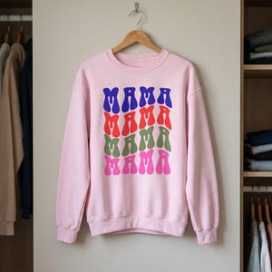 Puede incluir: Sudadera rosa claro con la palabra "MAMA" impresa en azul, rojo, verde y rosa, apilada verticalmente. La sudadera está colgada en una percha de madera frente a un armario.