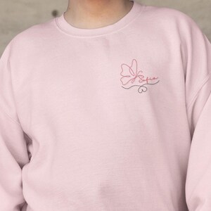 Puede incluir: Una sudadera rosa claro con un delicado diseño bordado. El diseño presenta una mariposa y el nombre "Sofia" en escritura cursiva. El bordado es en tonos rosa y negro.