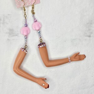 May include: Unique dangle earrings featuring doll arms with painted nails and bracelets. The earrings have pink seashell charms, purple and white beads, and gold-colored hooks. The earrings are attached to a pink glittery card.